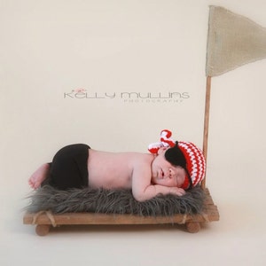 May include: A newborn baby wearing a red and white striped pirate hat with an eye patch, sleeping on a wooden platform with a gray fur rug. The platform has a small sail attached to it.