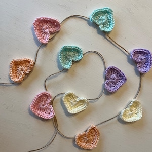 May include: A garland of crocheted hearts in various pastel colours, including pink, peach, yellow, light blue, and purple, strung together with twine.