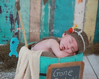May include: A newborn baby wearing a brown crocheted hat with a red and yellow fishing lure on top, sleeping in a turquoise barrel with a sign that reads "Gone fishin'" and a fishing net in the foreground.