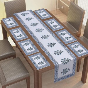 Può includere: Un runner da tavola bianco e otto tovagliette rettangolari abbinate con un motivo floreale blu stampato a blocchi e un bordo decorativo blu. Il set è disposto su un tavolo da pranzo in legno con sei sedie imbottite.