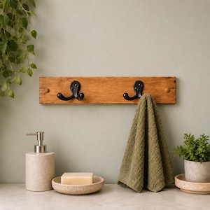 May include: A rectangular wooden wall rack featuring two black metal double hooks. The rack has a natural wood grain finish and is mounted horizontally. A textured olive green towel hangs from the right hook, adding a soft contrast to the wood.