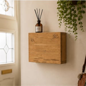 May include: A rectangular, light brown wooden wall-mounted shelf with a visible wood grain and knot pattern. A dark brown glass bottle containing black reed diffuser sticks sits on the top surface of the wooden shelf against a plain wall.