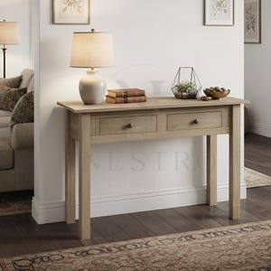 May include: A light brown wooden console table featuring two drawers with small dark knobs. The table surface holds a beige table lamp with a rounded base, two stacked brown books, a geometric wire terrarium, and a small bowl of stones.