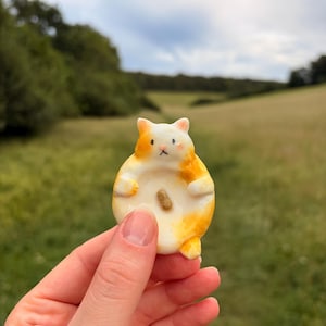 Può includere: Una piccola statuetta di gatto in ceramica, rotonda, con macchie bianche e arancioni. Il gatto presenta una pancia piatta e concava con un singolo punto marrone al centro, piccole orecchie, occhi dipinti, un naso minuscolo e una bocca.