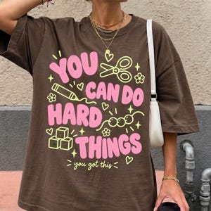 May include: A brown oversized t-shirt featuring a graphic print in pink and yellow. The text reads "YOU CAN DO HARD THINGS" and "you got this" with illustrations of scissors, a pencil, beads, and blocks. The shirt has short sleeves.