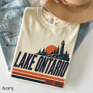 以下が含まれることがあります： 灯台、松の木、夕日のグラフィックが描かれたアイボリーのTシャツ。ダークブルーの文字で「LAKE ONTARIO EST. 1956」と書かれ、その下にオレンジとブルーの水平ストライプがあります。首元にはComfort Colorsのブランドラベルが付いています。