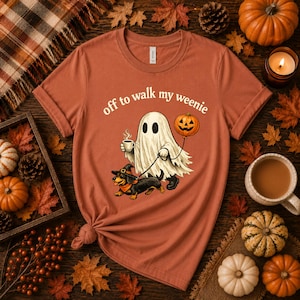 Pode incluir: Uma t-shirt cor de ferrugem com o gráfico de um fantasma segurando uma chávena de café e um balão de abóbora, a passear um cão salsicha com um chapéu de bruxa. O texto acima do gráfico diz, off to walk my weenie, em fonte branca.