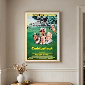 May include: A framed movie poster for Caddyshack featuring a green golf course, a large gopher, and portraits of four actors. The text reads Some People Just Don't Belong, Caddyshack, and lists the cast and crew in yellow and white lettering.