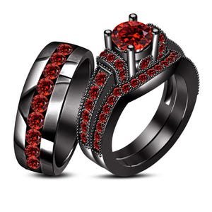 Könnte beinhalten: Ein zweiteiliges Ring-Set aus schwarzem Metall, besetzt mit leuchtend roten, rund geschliffenen Steinen. Der Verlobungsring zeigt einen markanten, zentralen roten Stein in einer Vier-Krappen-Fassung, umgeben von kleineren roten Steinen.