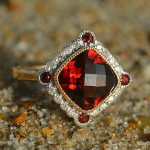 Könnte beinhalten: Ein Ring mit einem großen, quadratisch geschliffenen, tiefroten Granat-Edelstein in der Mitte, umgeben von einer gewellten Silberfassung, die mit kleinen weißen Steinen und vier kleineren runden roten Granaten an den Seiten besetzt ist.