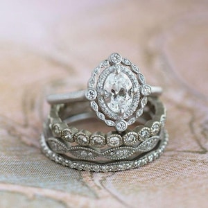 Könnte beinhalten: Ein Stapel aus drei silberfarbenen Ringen. Der oberste Ring hat einen oval geschliffenen Mittelstein, umgeben von einem Halo aus kleinen runden Steinen in einer gewellten Fassung. Der mittlere Ring hat eine Reihe runder Steine, der untere ist schmal.