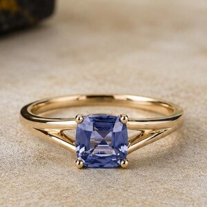 May include: A gold ring featuring a cushion-cut periwinkle blue gemstone set in a four-prong mounting. The band has a split shank design that tapers toward the center stone, creating a polished and refined appearance for this piece of jewelry.