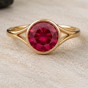 Peut inclure: Une bague en or ornée d'une pierre précieuse ronde, facettée et rouge profond, montée dans un serti clos. L'anneau de la bague se divise en deux bras fins en or poli qui se rejoignent au niveau de la pierre centrale pour un rendu élégant.