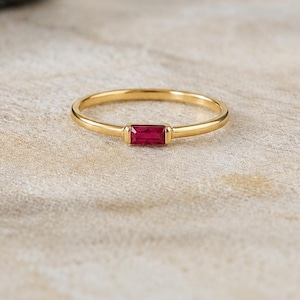 Op de afbeelding: Een dunne gouden ring met een enkele rechthoekige, in baguette geslepen rode edelsteen die horizontaal in het midden is gezet. De ringband is glad en gepolijst en toont een strak ontwerp met een levendige karmozijnrode steen als middelpunt.