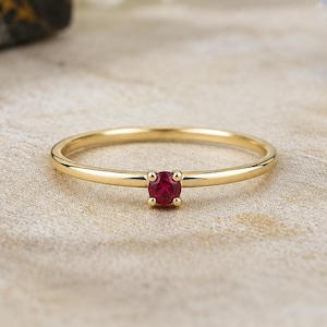 Peut inclure: Une fine bague en or avec une petite pierre précieuse rubis rouge de taille ronde, fixée dans un sertissage à quatre griffes. La bague présente une finition lisse et polie, mettant en valeur la couleur rouge vive de la pierre centrale.