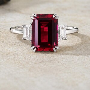 Puede incluir: Un anillo de plata con una gran piedra preciosa de color rojo intenso de corte esmeralda rectangular en el centro, flanqueada por dos piedras más pequeñas, claras y rectangulares de corte baguette a cada lado, en una banda de metal pulido.