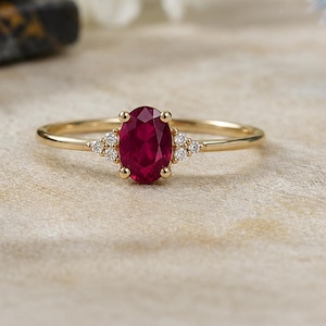 May include: A gold ring featuring a central oval-cut deep red ruby gemstone. The ruby is flanked on each side by three small, clear round-cut diamonds set in a cluster. The band is thin, polished gold, creating a delicate and refined appearance.