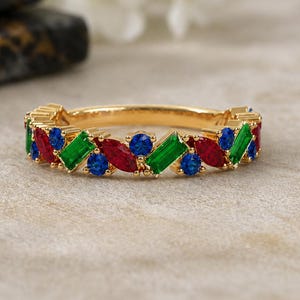 Pode incluir: Um anel de banda dourada com um padrão repetitivo de pedras vermelhas em corte marquise, pedras verdes em corte baguette e pedras azuis em corte redondo, cravadas numa fila contínua ao longo da circunferência exterior da banda.