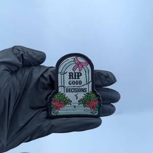 Op de afbeelding: Een geborduurde patch in de vorm van een grafsteen met een lichtblauwe achtergrond en een zwarte rand. De tekst RIP GOOD DECISIONS staat in zwarte letters. Bovenaan zit een roze strik en onderaan staan trossen rode rozen met groene bladeren.