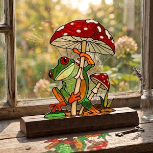Peut inclure: Un attrape-soleil en vitrail représentant une grenouille verte aux yeux rouges tenant un grand champignon rouge et blanc, accompagné d'un plus petit. L'objet est fixé sur un socle en bois rectangulaire et projette une ombre colorée sur la surface.