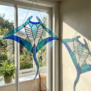 May include: A stained glass suncatcher shaped like a manta ray, featuring various shades of blue and teal glass pieces joined by dark metal solder lines. The piece is suspended by a thin metal chain and casts a colorful shadow on the wall.