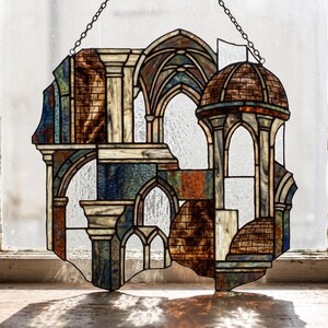 May include: A stained glass panel featuring an architectural scene with arched doorways, columns, and a domed structure. The piece uses textured glass in shades of brown, blue, and cream, held together by dark metal cames, suspended by a metal chain.