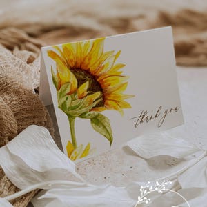 May include: A folded white card featuring a watercolor sunflower illustration. The sunflower is yellow and brown with green leaves. The words "thank you" are written in cursive. The card is from Ann Green Card Company.