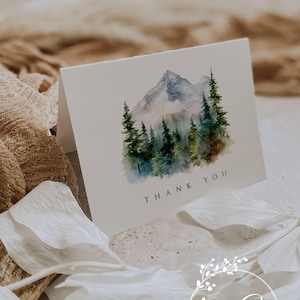May include: A folded white card with a watercolor painting of a mountain and evergreen trees. The words "THANK YOU" are printed in black. The card is part of a stationery set, with the designer's logo at the bottom.