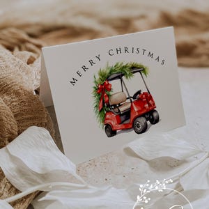 May include: A folded white Christmas card featuring a watercolor illustration of a red golf cart decorated with a green wreath and red bow. The words "MERRY CHRISTMAS" are printed at the top. The card is branded with "Ann Green DESIGNS - CARDS".