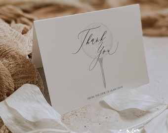 May include: A folded white card with the words "Thank You" in elegant script, featuring a golf ball design. The text "FROM THE DESK OF ALAN GRAY" is below the main text. The card is surrounded by white floral elements and a beige fabric.