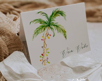 May include: A folded white greeting card featuring a watercolor illustration of a palm tree decorated with colorful lights. The words "Warm Wishes" are written in cursive. The Ann Green Card Company logo is at the bottom.