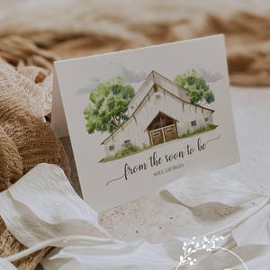 May include: A folded white card with a watercolor illustration of a barn, trees, and text that reads "from the soon to be MRS. GEORGIA". The card is surrounded by white floral accents and a beige fabric.
