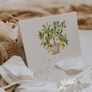 May include: A white folded greeting card featuring a watercolor illustration of a lemon tree in a decorative patterned pot. The word GRAZIE is printed in black capital letters below the illustration. The card has a clean, simple design.