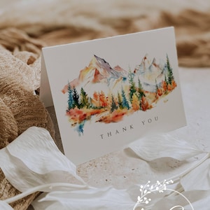 May include: A folded white card with a watercolor painting of a mountain range, trees, and autumn foliage. The words "THANK YOU" are printed in black. The card is decorated with a floral logo that says "Ann Green Designs - Cards".