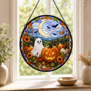 Może przedstawiać: Okrągły witrażowy łapacz słońca przedstawiający scenę halloweenową ze świecącą dynią, białym duchem, pomarańczowymi dyniami i słonecznikami. W tle widoczny jest sierp księżyca, nietoperze oraz drewniany płot pod niebieskim niebem.