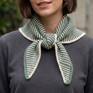 Puede incluir: Una bufanda triangular de punto con textura acanalada y rayas alternas en color verde salvia y crema. La bufanda está atada con un nudo sencillo en la parte delantera y tiene un borde liso de color crema a lo largo de los extremos.