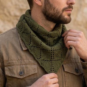 Puede incluir: Un pañuelo triangular de punto en color verde oliva con un patrón de encaje de rombos repetitivo y pequeños detalles de ojales. El pañuelo se lleva alrededor del cuello sobre una chaqueta beige con botones, dos bolsillos en el pecho y puños.
