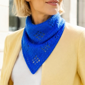 May include: A vibrant royal blue triangular knit scarf featuring a delicate openwork diamond pattern. The scarf is worn around the neck, layered over a plain white top and a light yellow blazer, showcasing its textured, lightweight construction.