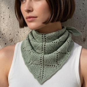 Op de afbeelding: Een saliegroene driehoekige gebreide bandana met een ingewikkeld kantpatroon met oogjesdetails en een geribbelde textuur. Het accessoire is in de nek geknoopt en wordt gedragen over een eenvoudig wit mouwloos topje met een ronde halslijn.