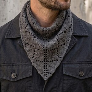 May include: A charcoal gray triangular knit scarf featuring an intricate openwork lace pattern with a pointed bottom edge. The scarf is worn around the neck over a dark charcoal button-down jacket with two chest pockets and visible buttons.