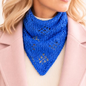 May include: A vibrant blue knitted triangular scarf featuring a delicate openwork diamond pattern throughout the fabric. The scarf is worn around the neck, layered over a white ribbed top and a light pink coat with wide lapels and structured shoulders.