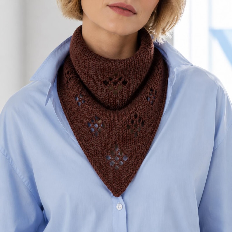 May include: A dark brown knitted neck warmer featuring a triangular shape and a series of diamond-patterned eyelet cutouts. The accessory is worn around the neck over a light blue collared button-down shirt, showcasing a textured yarn construction.