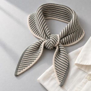 Puede incluir: Un pañuelo de punto con un patrón de rayas gris oscuro y blanco roto. El accesorio tiene un borde liso en color blanco roto y está atado con un nudo sencillo, descansando junto a una camisa blanca de cuello con un botón visible.
