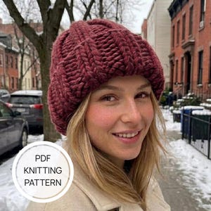 May include: A chunky knit beanie in a deep burgundy color featuring a thick ribbed texture. The hat has a rounded crown and a wide folded brim. A circular white graphic overlay on the image contains the text PDF KNITTING PATTERN.