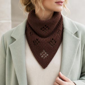 May include: A dark brown knitted triangular scarf featuring a ribbed texture and a series of openwork diamond-shaped patterns. The scarf is worn around the neck, layered over a light-coloured top and a pale green coat with structured lapels.