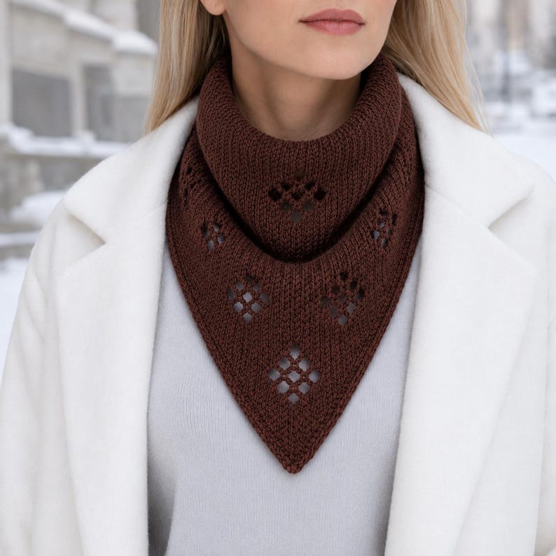 May include: A dark brown knitted triangular scarf featuring a ribbed texture and a series of diamond-shaped eyelet cutouts arranged in a pattern. The scarf is worn around the neck, layered over a light grey top and a white coat.
