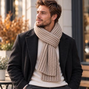 May include: A beige ribbed knit scarf worn over a white sweater and a dark charcoal wool coat. The scarf features a thick, textured vertical ribbed pattern that wraps around the neck, providing a warm and layered look for cold weather attire.