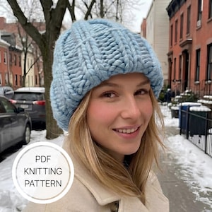 May include: A light blue chunky knit beanie with a ribbed texture. The hat features a thick, soft yarn construction with a folded brim. A circular white graphic overlay on the lower left corner contains the text PDF KNITTING PATTERN.