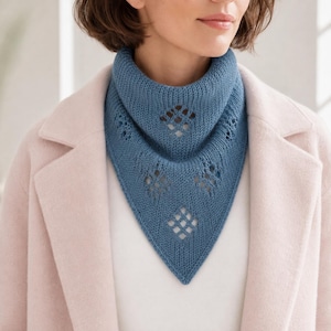 May include: A blue knitted triangular scarf featuring a ribbed texture and a series of diamond-shaped eyelet cutouts arranged in a vertical pattern down the center. The scarf is worn around the neck over a white shirt and a light pink coat.