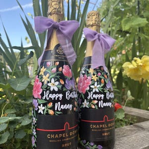 May include: Two bottles of Chapel Down Brut English sparkling wine, each featuring a hand-painted floral design in pink, purple, and yellow, with the text Happy Birthday Nanny in white script and a light purple ribbon tied around the bottle neck.